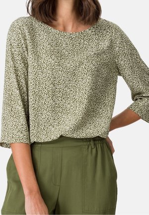 Frau trägt eine grün-weiß gemusterte Bluse mit dreiviertel Ärmeln und festliche, hoch taillierte Olivgrüne Hose.