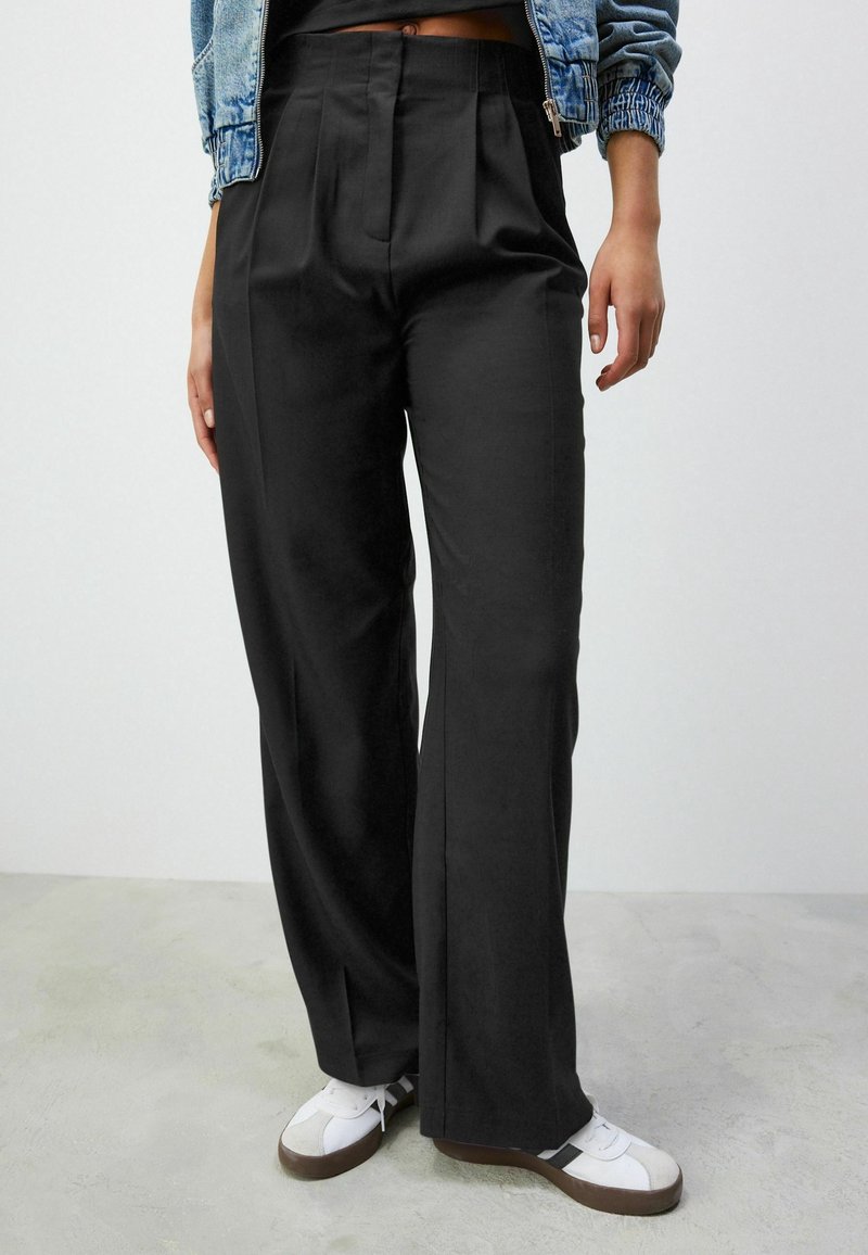 Pantalon noir à jambes larges avec plis, tissu lisse, taille haute et poches latérales, assorti à des baskets blanches avec des accents marron.