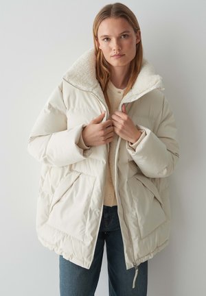 Giacca bianca gonfiabile con un ampio colletto morbido color crema, due tasche davanti e chiusura con zip, indossata sopra un maglione beige e jeans blu.