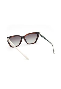 Gafas de sol de estilo ojo de gato con montura de carey marrón, lentes grises degradados y patillas blancas que presentan un patrón geométrico en verde y negro.