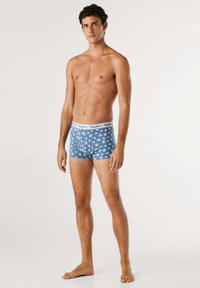 Blaue Boxer-Shorts mit einem weißen Bündchen, das das Markenlogo trägt, geschmückt mit einem sich wiederholenden weißen Blumenmuster.