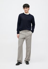 Marinegenser, hvit skjorte innvendig og lysegrå stripebukser. Svarte sko. Modellen står foran en nøytral bakgrunn, med hendene i lommene.