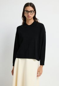 Schwarzer, gestrickter Pullover mit großem Kragen und Perlendetails. Lange Ärmel und lockere Passform, kombiniert mit einem cremfarbenen Rock.