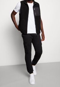 Muž v černé fleecové vestě přes bílé tričko, černých cargo kalhotách a bílých teniskách, stojící na pozadí jednoduchého světlého pozadí.