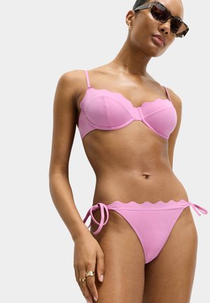Kvinde iført lyserød bikini med bølget kant og bindesidebunde samt skildpaddeskal-solbriller på hvid baggrund.