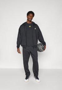 Schwarzes Sportoutfit bestehend aus einer Kapuzenjacke, einem T-Shirt und Jogginghosen. Mit gelben Logodetails. Hält einen schwarzen Basketball.