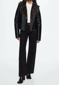 Veste en cuir noir avec col en fausse fourrure grise, fermeture éclair, portée sur un haut blanc et un jean noir évasé, assortie à des bottines noires.