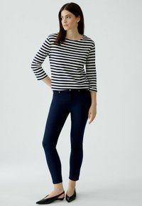 Gestreepte lange mouwen top in marineblauw en wit, gecombineerd met donkerblauwe skinny jeans. Model draagt zwarte platte schoenen en staat tegen een lichte achtergrond.