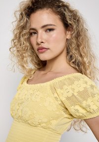 Een jonge vrouw met krullend haar poseert zelfverzekerd in een lichtgele blouse met bloemenpatronen, en toont een natuurlijke make-uplook en een ernstige uitdrukking.