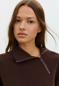 Hnědý zip-up pulovr s malým límečkem, vyrobený z hladkého materiálu. Obsahuje stříbrný zip a ploché švy pro čistý vzhled.