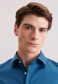 Niebieska koszula z długim rękawem, zapinana na guziki, z kołnierzykiem typu spread, wykonana z gładkiej tkaniny, w dopasowanym kroju, z białymi guzikami na listwie zapięcia.