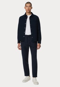 Camicia navy con bottoni e due tasche sul petto, t-shirt bianca sotto, pantaloni navy con cordino e sneakers bianche. Tessuto dalla consistenza liscia.