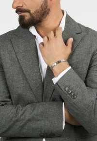 Blazer grigio a scacchi con maniche a due bottoni, indossato sopra una camicia bianca, che mette in mostra un braccialetto argento con un accento nero.