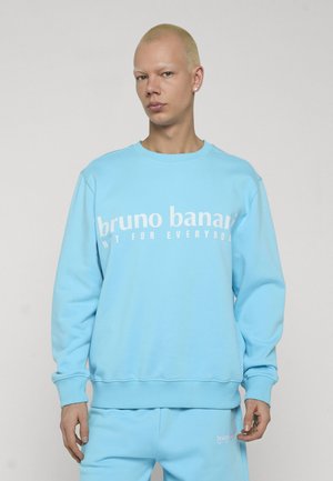 Persoon draagt lichtblauwe sweatpak met bijpassende broek met witte tekst "bruno banani", staand tegen een effen lichte achtergrond.