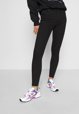 Osoba ubrana w czarne, dopasowane spodnie i białe sneakersy New Balance z fioletowymi i turkusowymi akcentami, stojąca na tle jednolitego tła.