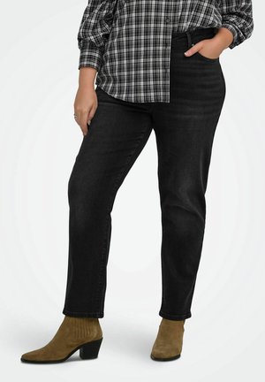 Personne portant un pantalon noir, des bottines en daim beige et une chemise à carreaux noir et blanc, une main dans la poche.