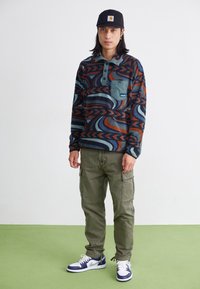 Ung person iklädd en svart keps, mönstrad långärmad tröja, olivgröna cargobyxor och vit-blå sneakers som står på ett grönt golv.