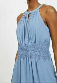Robe bleu clair avec un corsage plissé, un col halter et des détails en dentelle délicats à la taille. Tissu lisse avec une silhouette fluide.