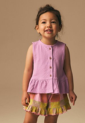 Jeune fille aux cheveux foncés souriante, portant un haut rose sans manches à boutons et un short coloré à volants, sur un fond beige uni.
