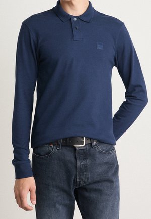 Navy long-sleeve polo shirt made of cotton, featuring a collar, two buttons, and a small brand logo patch on the chest. Paired with dark jeans.