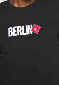 Camiseta negra con texto blanco "BERLIN" y un logo cuadrado rojo de un jugador de baloncesto en el aire a la derecha del texto.