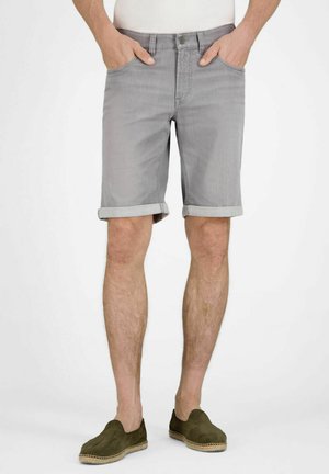 Man die lichtgrijze shorts met opgerolde zoom, wit overhemd en olijfgroene instapschoenen draagt, met handen in de zakken staand.