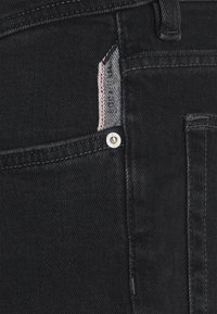 Vaqueros de mezclilla negra con un bolsillo frontal que presenta un detalle a rayas rojas y blancas, herrajes plateados y un diseño minimalista.
