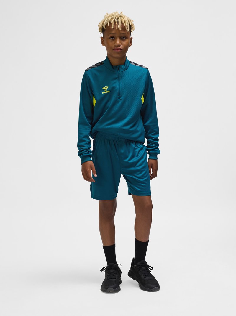 Garçon avec des dreadlocks blonds portant un survêtement de sport bleu sarcelle avec des accents jaunes, des chaussettes noires et des chaussures de sport noires, debout sur un fond gris.