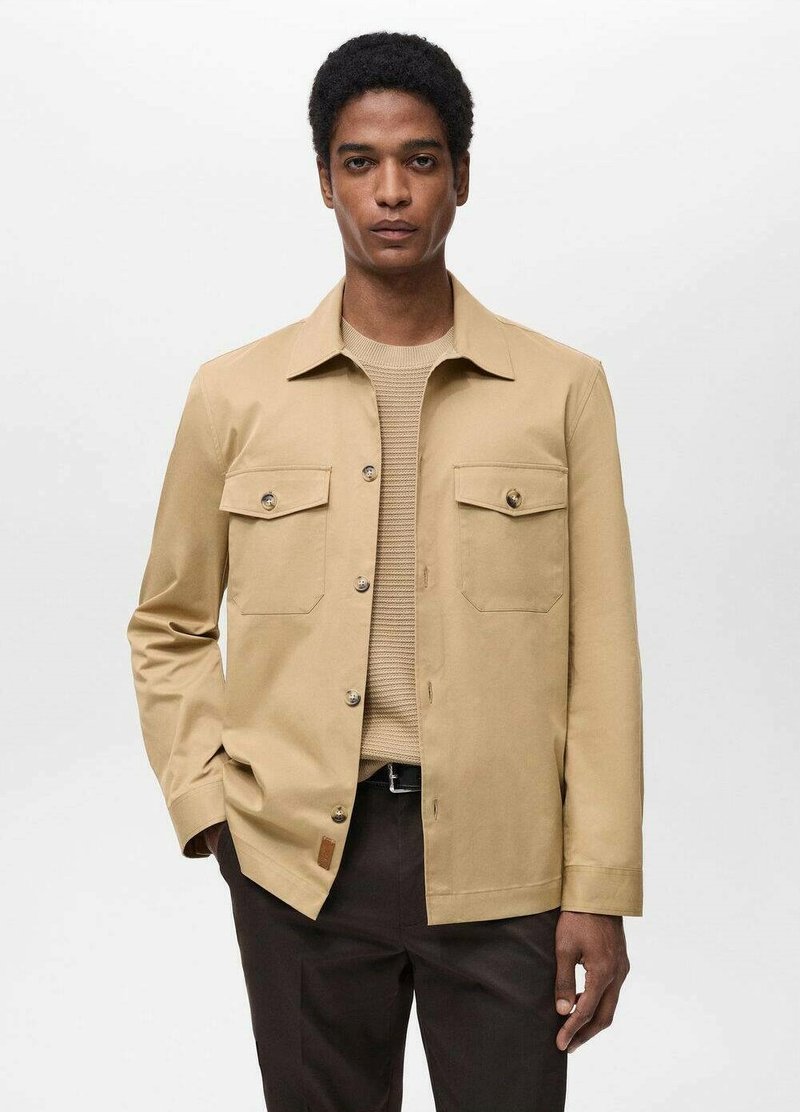 Lys beige sweater med struktur iført en tan-jakke med krave, to påsyede lommer, frontknapper og lange ærmer.