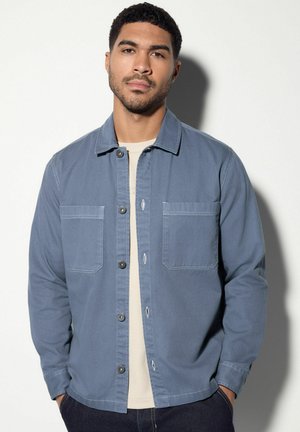 Cămașă din denim albastru cu două buzunare la piept, închidere cu nasturi și o croială lejeră, purtată peste un top rib knit de culoare deschisă.