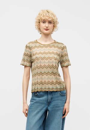 Ung person med krøllet blondt hår iført en kortærmet top med zigzag-mønster i beige, brun og lyseblå samt blå jeans mod en hvid baggrund.