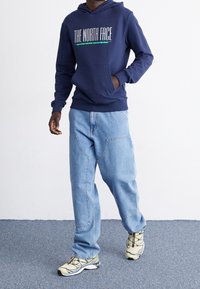 Marinblå hoodie med "THE NORTH FACE"-logotyp, matchad med ljusblå löst sittande jeans och flerfärgade sportskor.