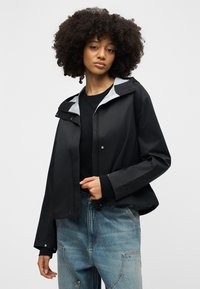 Γυναίκα με σγουρά μαλλιά που φοράει μαύρο σακάκι πάνω από μαύρο τοπ, σε συνδυασμό με ανοιχτό μπλε τζιν, στέκεται μπροστά σε απλό ανοιχτό φόντο.