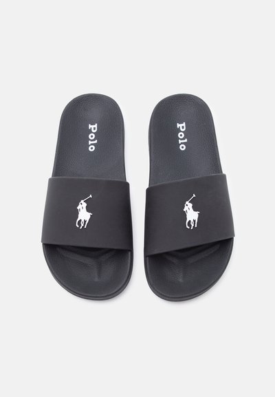 Μαύρα σανδάλια χωρίς λουράκια με άσπρο κείμενο "Polo" στο εσωτερικό και λευκό λογότυπο παίκτη πόλο σε φαρδιές λουρίδες, εμφανιζόμενα σε λευκό φόντο.