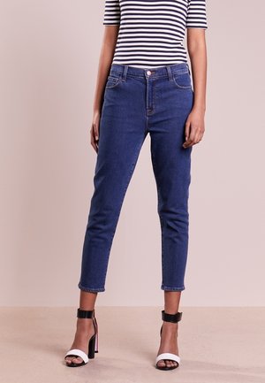 Kvinde iført blå skinny jeans med høj talje, sort-hvid stribet top og sort-hvide højhælede sandaler, der står på beige gulv.