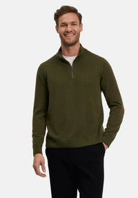 Maglione verde oliva con zip e collo alto, realizzato in tessuto morbido, con un design aderente, polsini e orlo a costine, indossato su una t-shirt bianca.