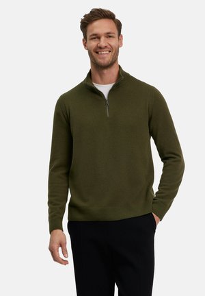 Olivengrøn zip-neck sweater lavet af blødt stof, med en tætsiddende design, ribbede manchetter og en kant, båret over en hvid t-shirt.