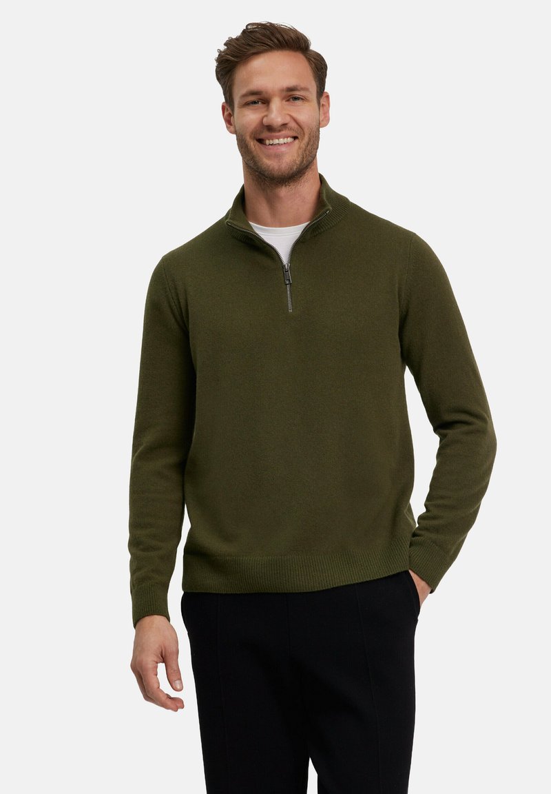 Maglione verde oliva con zip e collo alto, realizzato in tessuto morbido, con un design aderente, polsini e orlo a costine, indossato su una t-shirt bianca.