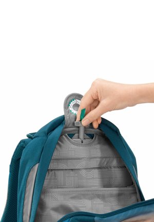 Teal fabric backpack interior with a hand adjusting a gray fastener. The lining features a geometric pattern in light gray.