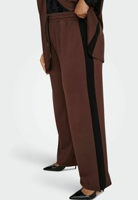 Osoba nosi smešane široke hlače smeđe boje s crnim bočnim trakama i crnim cipelama s oštrim prstima, ruke su joj na struku.