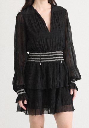 Femme portant une robe longue noire transparente à manches longues avec un décolleté en V profond, une ceinture et des détails aux poignets à motif zigzag blanc.