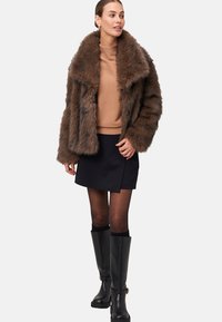 Veste en fausse fourrure marron avec un large col, portée sur un pull beige et une jupe noire. Des bottes noires hautes jusqu'aux genoux complètent la tenue.