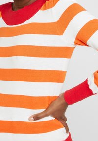 Pull rayé orange et blanc, avec un col et des poignets rouges. Texture tricotée douce, coupe ajustée et accents de couleur contrastants.