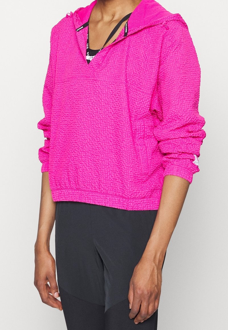 Pull de couleur rose vif, texturé, avec une capuche, doté d'un col en V et de poignets froncés. Adapté pour le sport, à porter avec un legging noir.