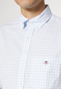 Muž v košili s jemným modrým a bílým kostkovaným vzorem, s knoflíky a malým štítkovým logem na kapsičce.