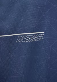 Close-up of navy blue fabric with subtle geometric pattern and white Hummel brand logo and stripe across the center.