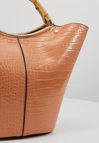 Tan handtas van krokodillenleer met een brede opening, glanzende afwerking en een goudkleurig rond handvat. Heeft een detail met een centrale naad.