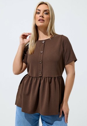 Kobieta z długimi blond włosami, ubrana w brązową bluzkę z krótkim rękawem, zapinaną z przodu na guziki z marszczoną talią, w zestawieniu z jasnoniebieskimi dżinsami.