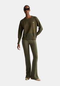 Maglione a maglia verde oliva con dettagli a costine, abbinato a pantaloni aderenti verde oliva con cuciture e bottoni decorativi in fondo.