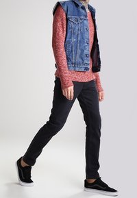 Denimväst över en röd texturerad långärmad skjorta, svarta slim-fit jeans och svarta sneakers. Outfiten har lagerstilar och avslappnad kl ädsel.